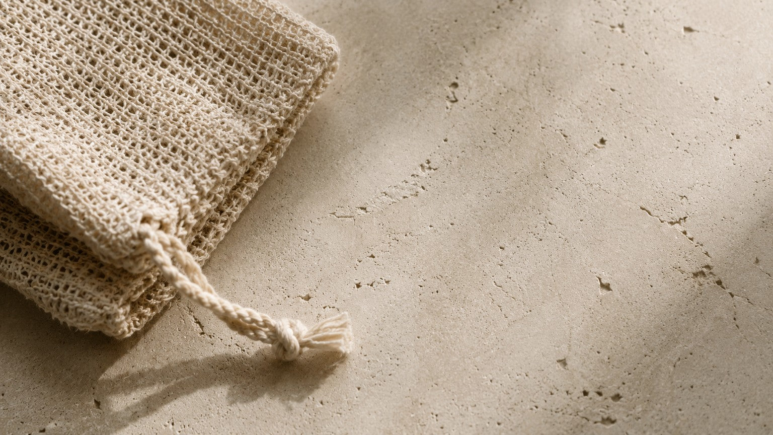 A folded natural beige burlap drawstring pouch with a knotted cord, placed on a textured concrete surface with soft shadows.