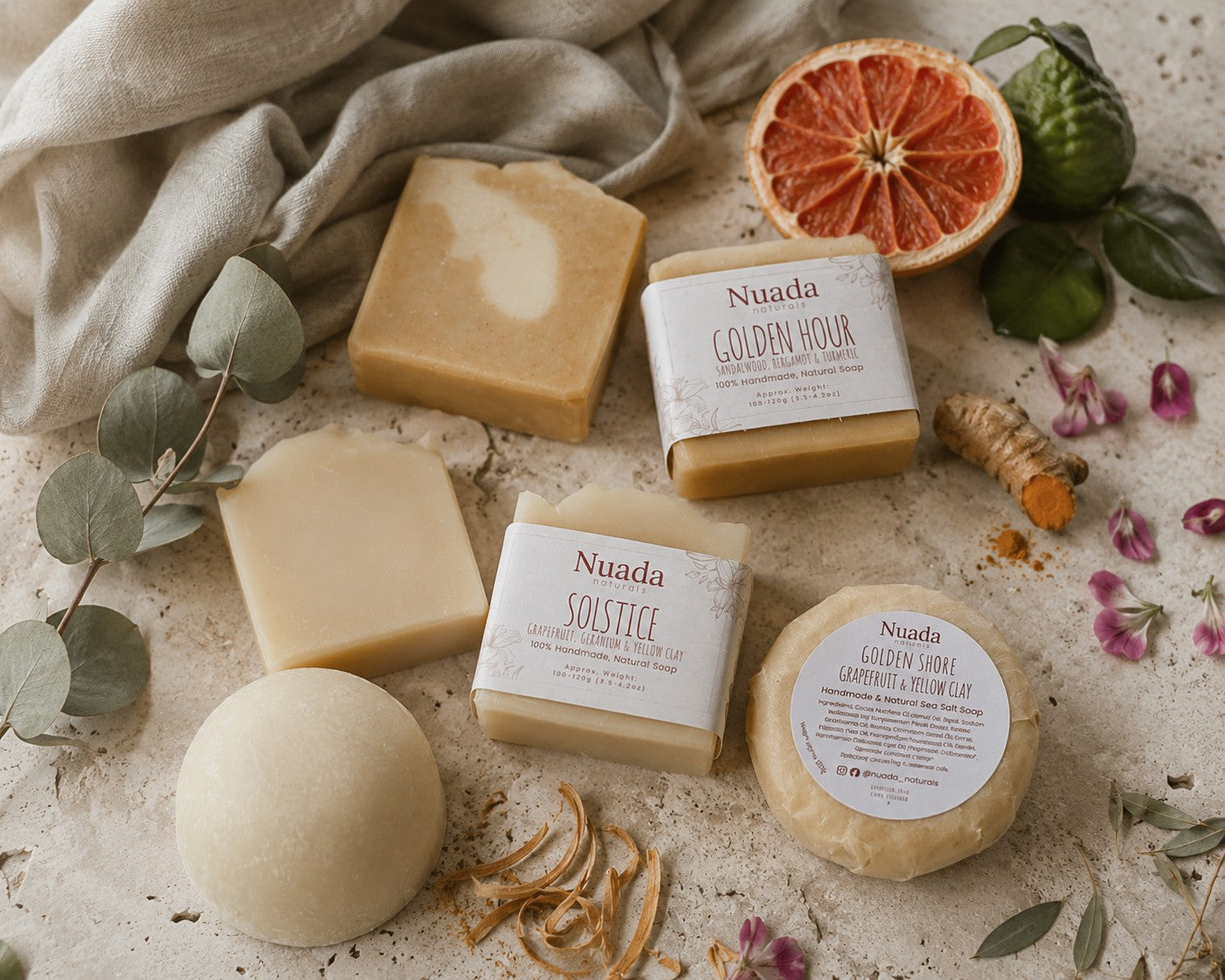 Four Nuada handmade soaps in various shapes and sizes, styled in a flat-lay arrangement with eucalyptus leaves, a grapefruit half, turmeric root, and a beige linen cloth on a textured stone surface.
