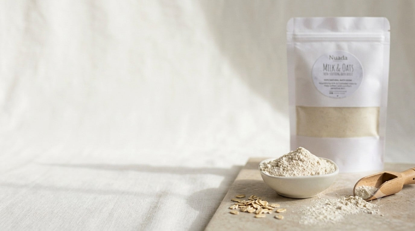 A stand-up pouch of Nuada Naturals Milk & Oats powder rests on a light surface beside a small bowl containing the powder, scattered oats, and a wooden scoop, against a soft, neutral fabric background.