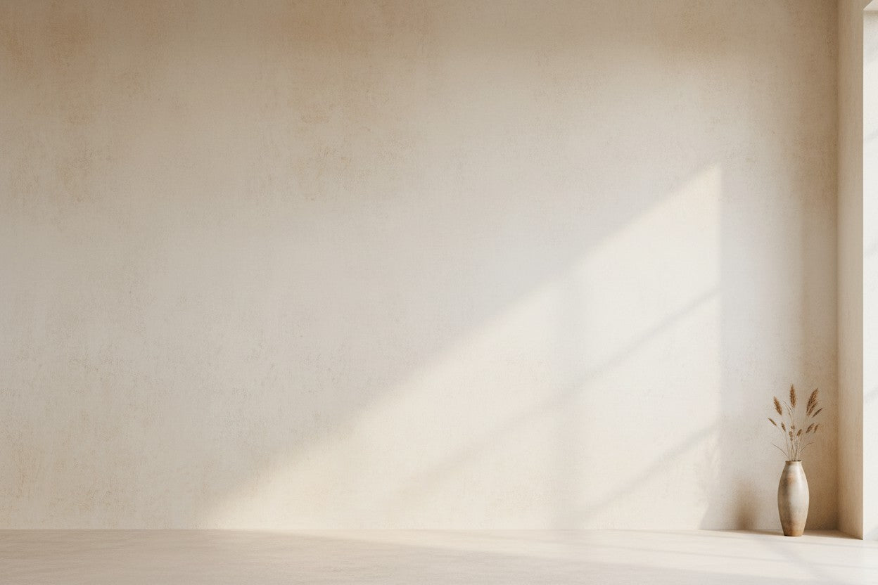 Minimalist beige room corner with textured plaster walls, soft sunlight casting diagonal shadows, and a small ceramic vase with dried stems on the floor near the right edge.